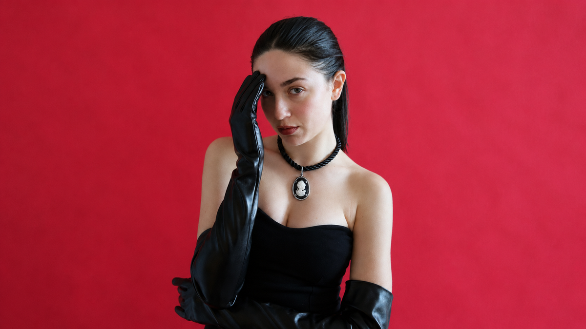 A woman with dark hair tied back wears a black strapless top, long black gloves, and a black choker with a cameo-style pendant, posed against a solid red background.