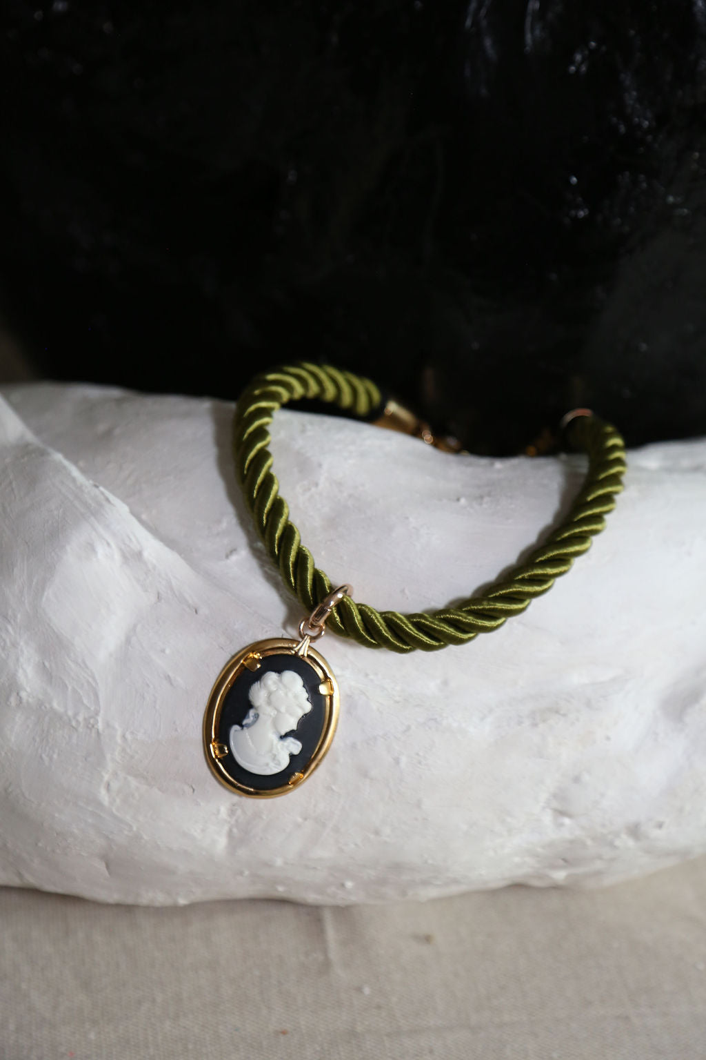 A twisted olive green and black cord necklace with a gold-tone oval cameo pendant, displayed on a white textured surface against a dark background.